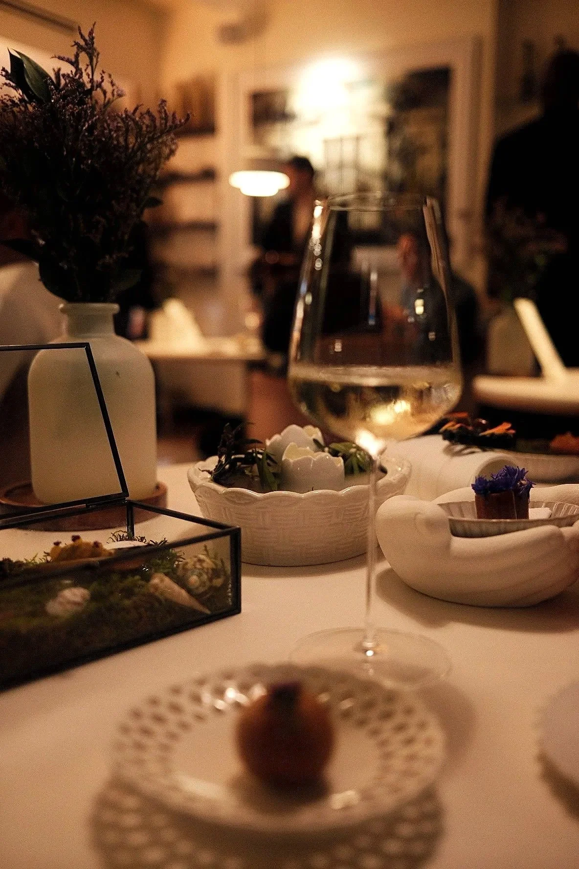 A table set with a glass of white wine, small decorative bowls with food or flowers, and a vase with flowers in a softly lit room, with people in the background.