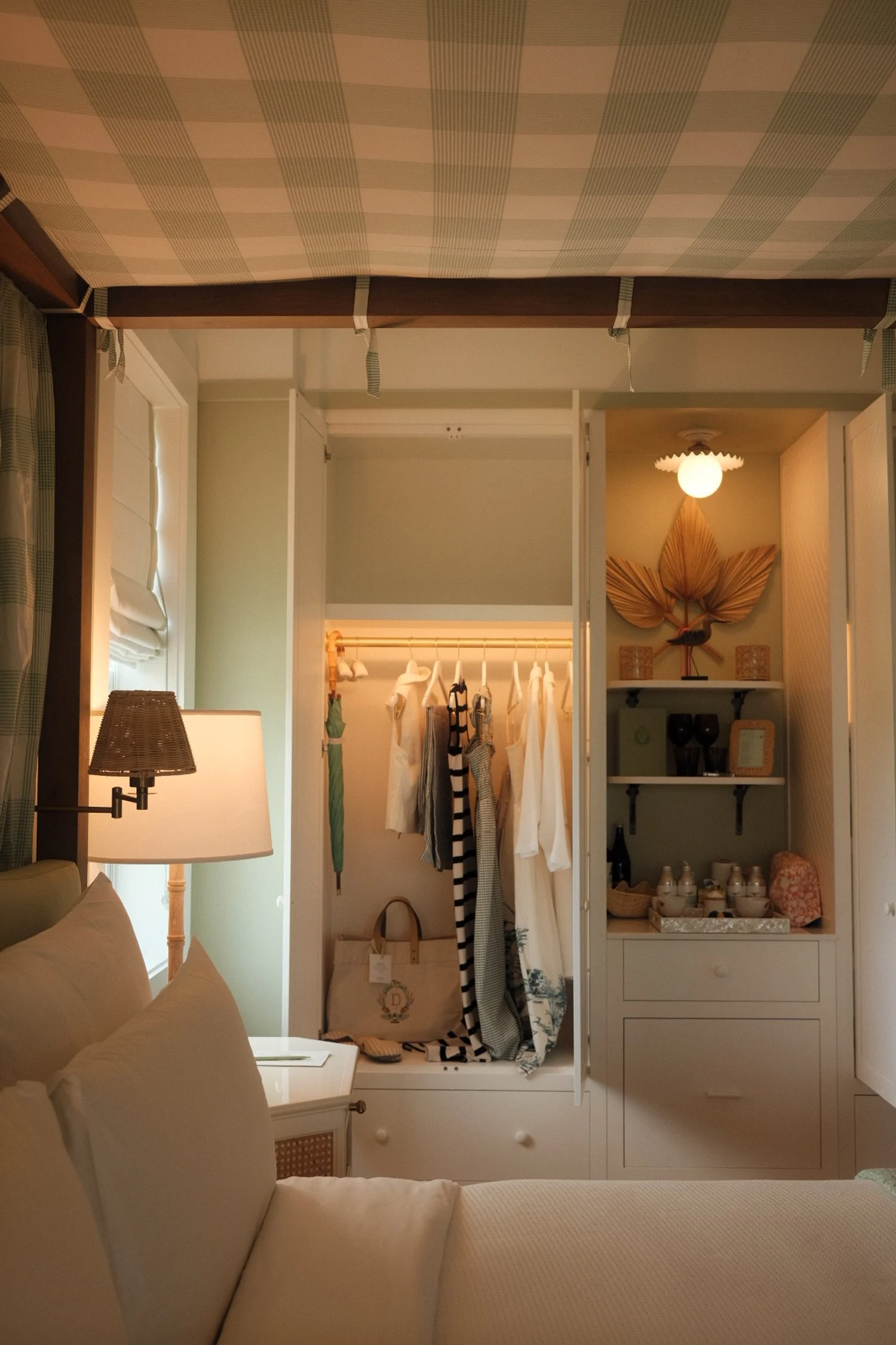 Interior view of a bedroom with an open closet revealing hanging clothes, a suitcase, and decorative shelves with decor and dishes.