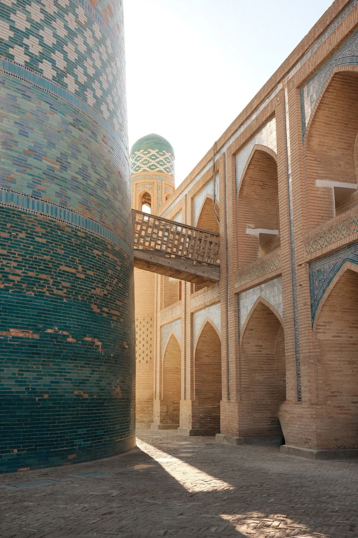 View of a historic Islamic courtyard with brick and tile architecture, arched doorways, a wooden bridge, and a blue-tiled minaret in the background.