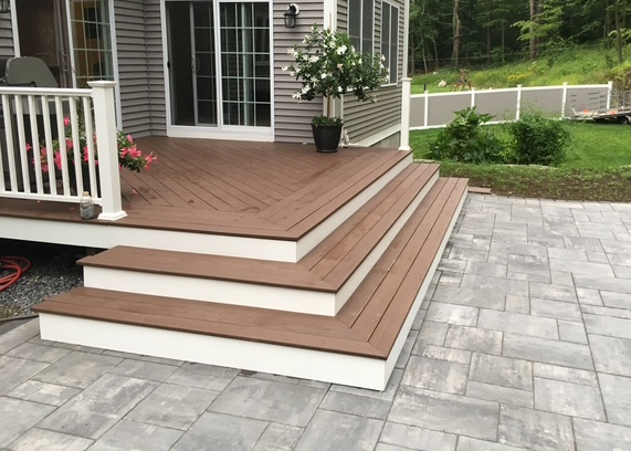Wooden deck with three steps leading to a house with large glass door, white railing, potted plant, and outdoor landscaping with green grass and trees.