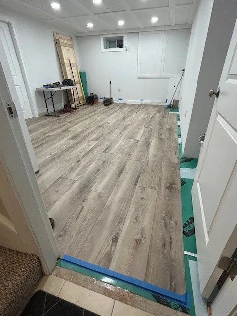Room under renovation with new wood laminate flooring, tools, and construction materials; door open to a small room with a table, chairs, and a window.