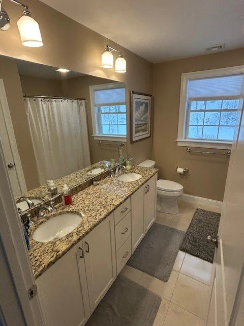 Bathroom with double sink granite countertop, white cabinets, two windows, a toilet, a bathtub with shower curtain, and gray rugs.