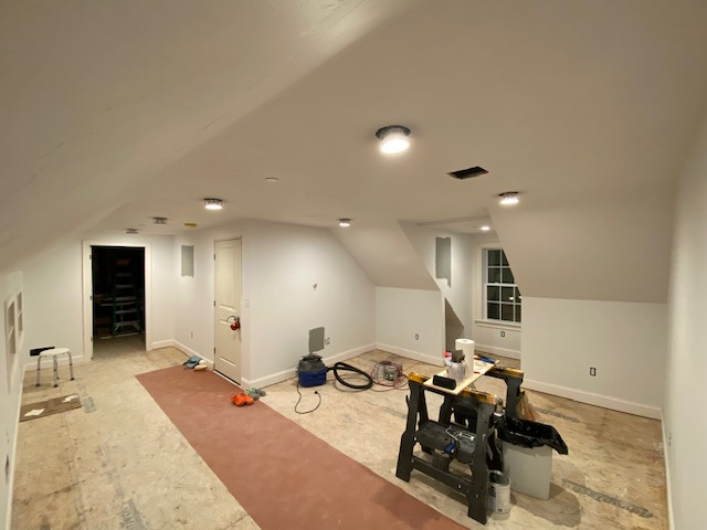 Empty attic room under renovation with tools, a workbench, and a window, partially carpeted floor.