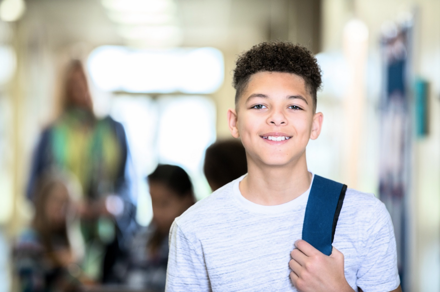 A smiling young boy with curly hair holding a backpack strap in a school hallway, with blurred students and a teacher in the background.