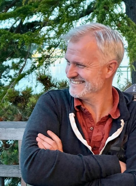 A smiling middle-aged man with gray hair and a beard sitting outdoors in front of green trees and a body of water, wearing a black jacket and a reddish-brown shirt.