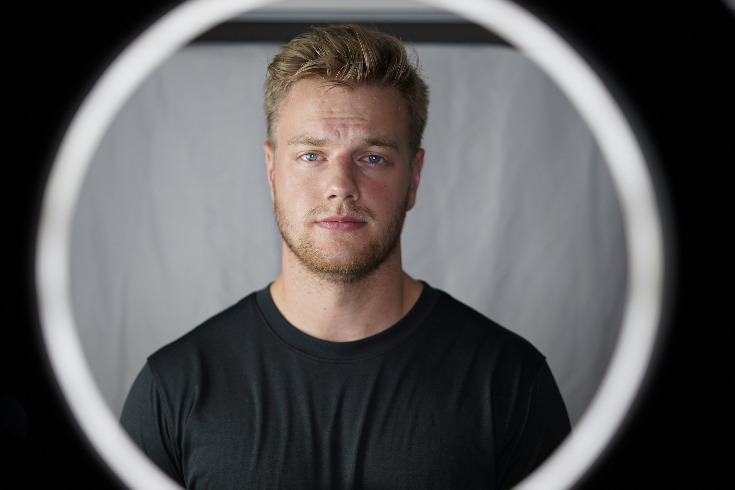 Portrait of a young man with short blond hair and blue eyes, wearing a black t-shirt, looking directly at the camera against a plain gray background, framed by a circular object.