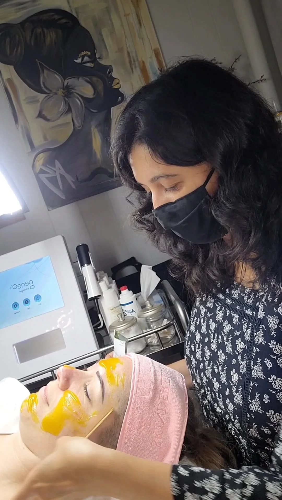 A woman receiving a facial treatment at a spa or clinic, lying on a bed with yellow facial mask applied, wearing a pink headband, while a cosmetologist in a black face mask applies or monitors the treatment.