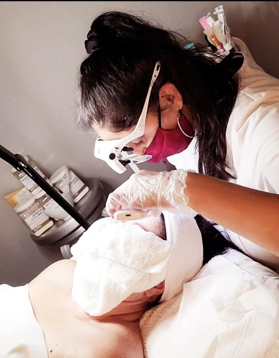 A woman wearing protective glasses, a face mask, and gloves is performing a cosmetic or medical procedure on a patient's face, who is lying down with their eyes covered.