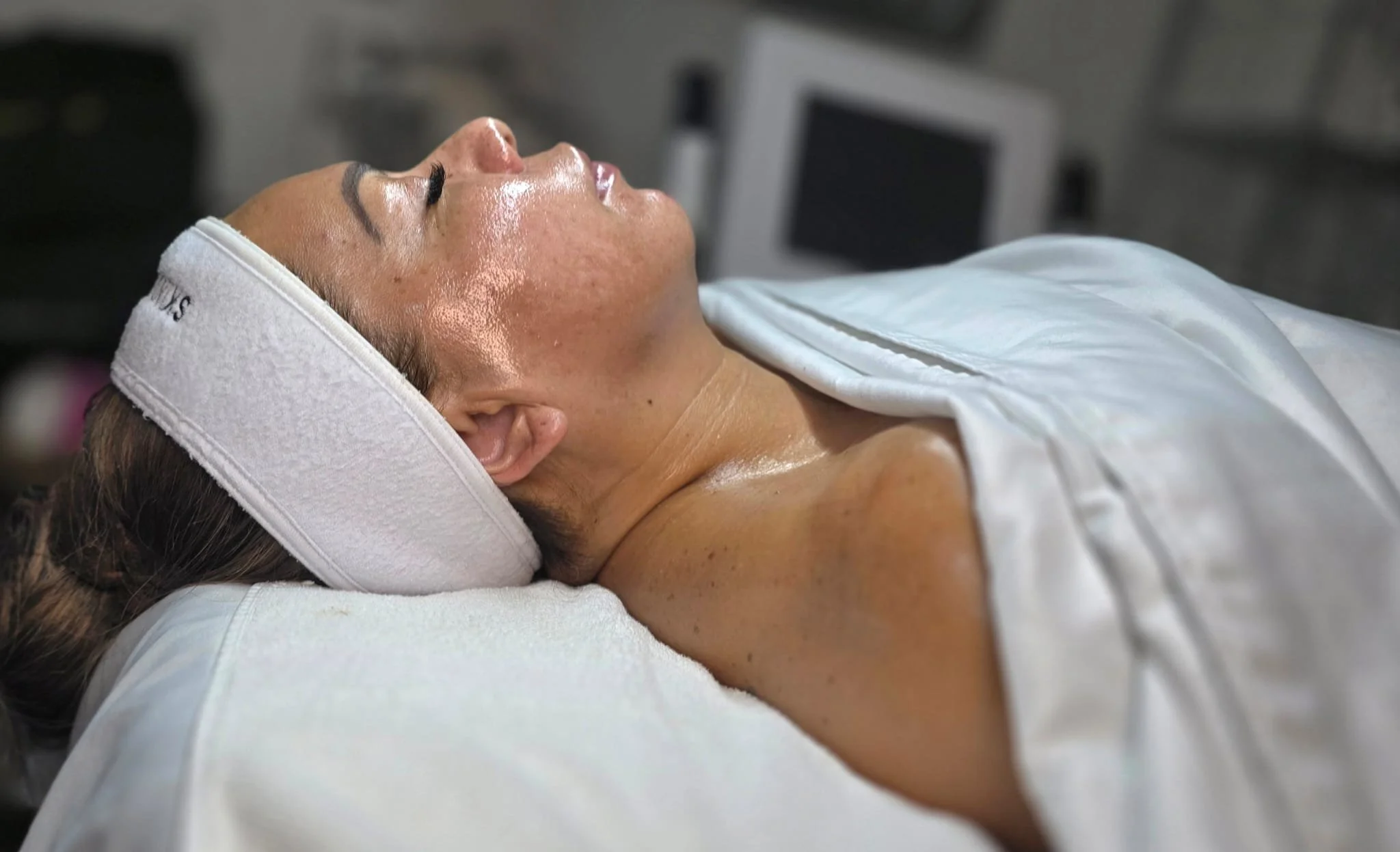 A woman lying on a treatment bed with her eyes closed, wearing a white towel and a white headband, in a spa or skincare clinic.