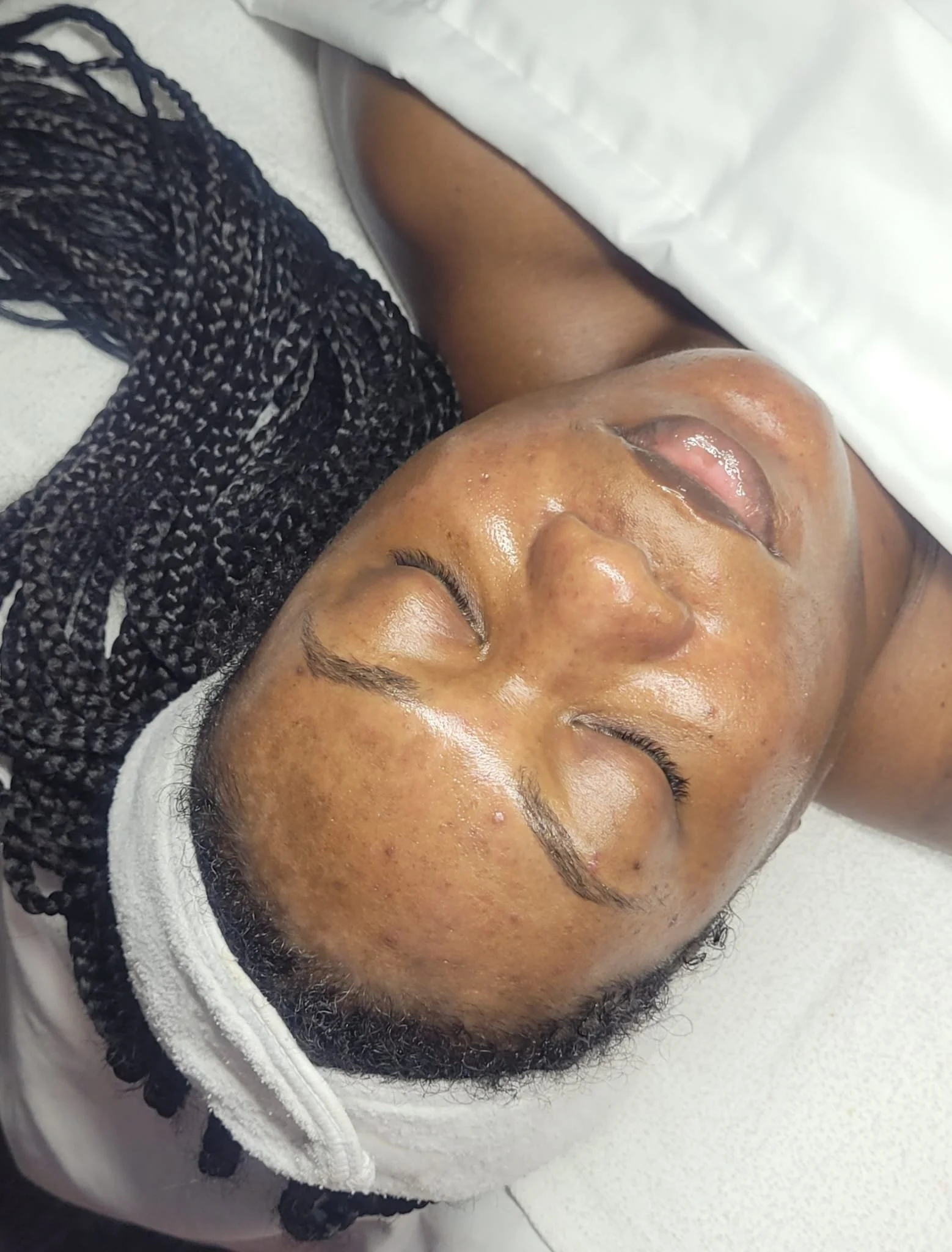 A woman lying on a treatment bed with a towel wrapped around her head, her eyes closed, receiving a facial skincare treatment.