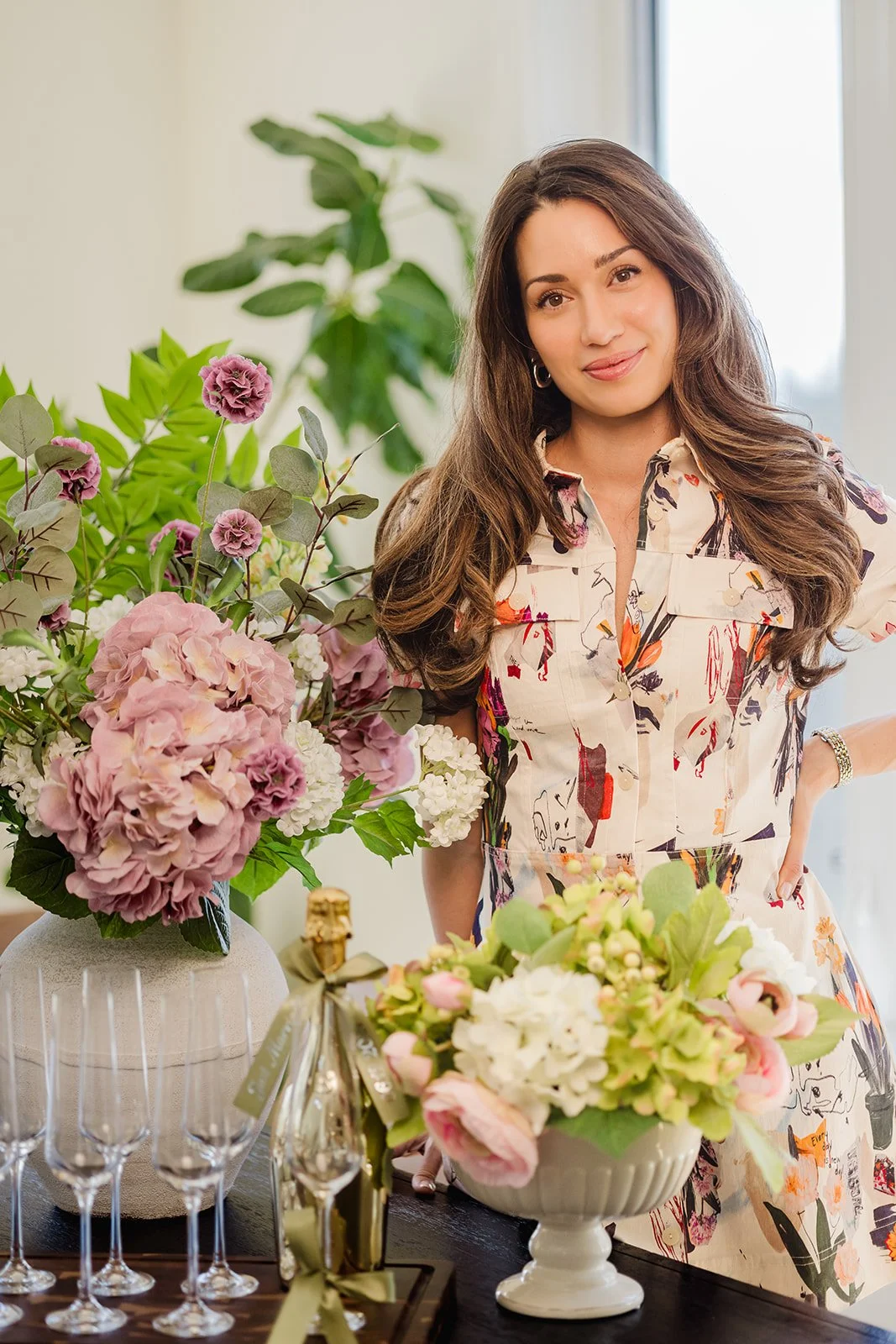 A woman with long, wavy brown hair and hoop earrings stands next to a large bouquet of pink, white, and purple flowers, wearing a colorful, patterned dress. The setting appears to be a bright indoor space with a window and green plant in the background.