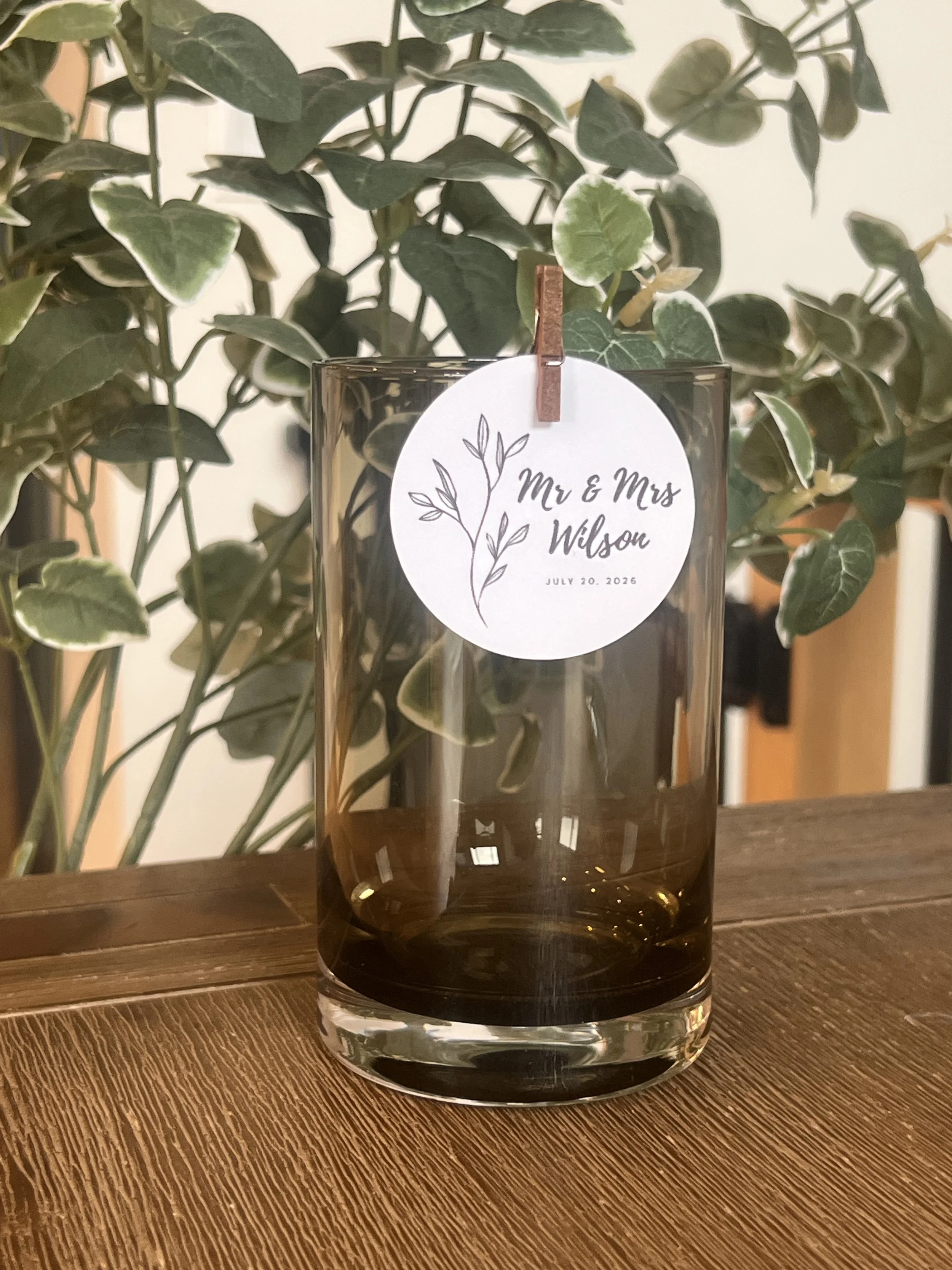 A glass vase with a label reading 'Mr & Mrs Wilson, July 20, 2026' holding a plant, placed on a wooden surface with a background of green foliage.
