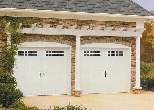 View of a two-car garage with white doors and brick accents, attached to a house with a gray shingle roof.