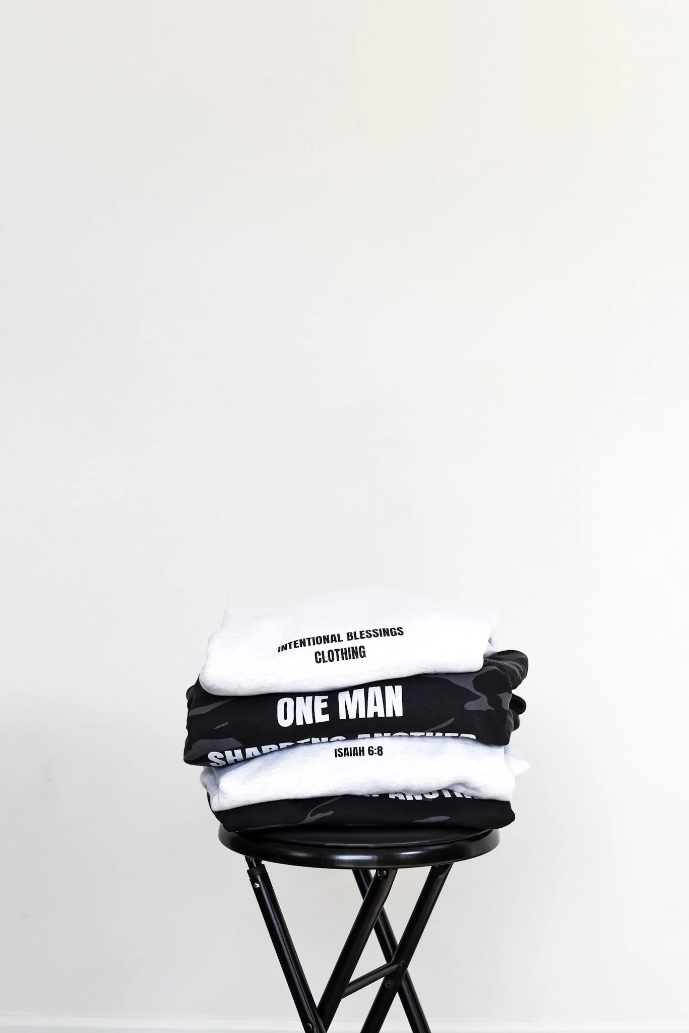 A stack of folded clothing, including shirts and hoodies with religious messages, on a black stool against a plain white wall.