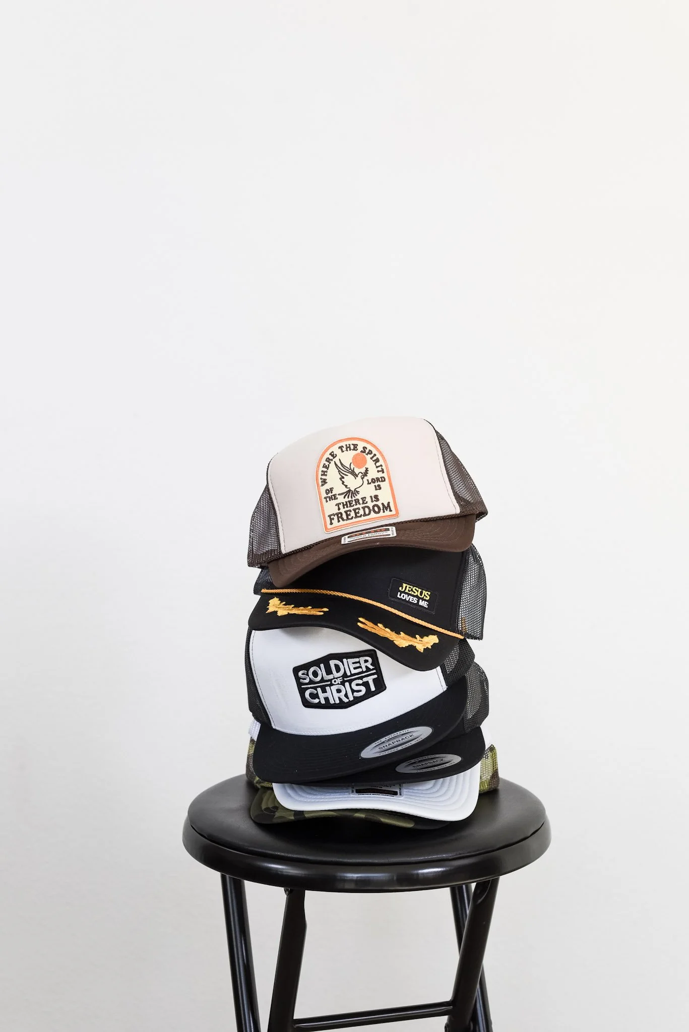 Stacked hats on a black stool, including a white and brown trucker hat with a patch that reads 'WHERE THE SPIRIT OF THE LORD IS, THERE IS FREEDOM', a black cap with yellow embroidery, a white cap with black embroidery reading 'SOLDIER OF CHRIST', and camouflage and white caps at the bottom.
