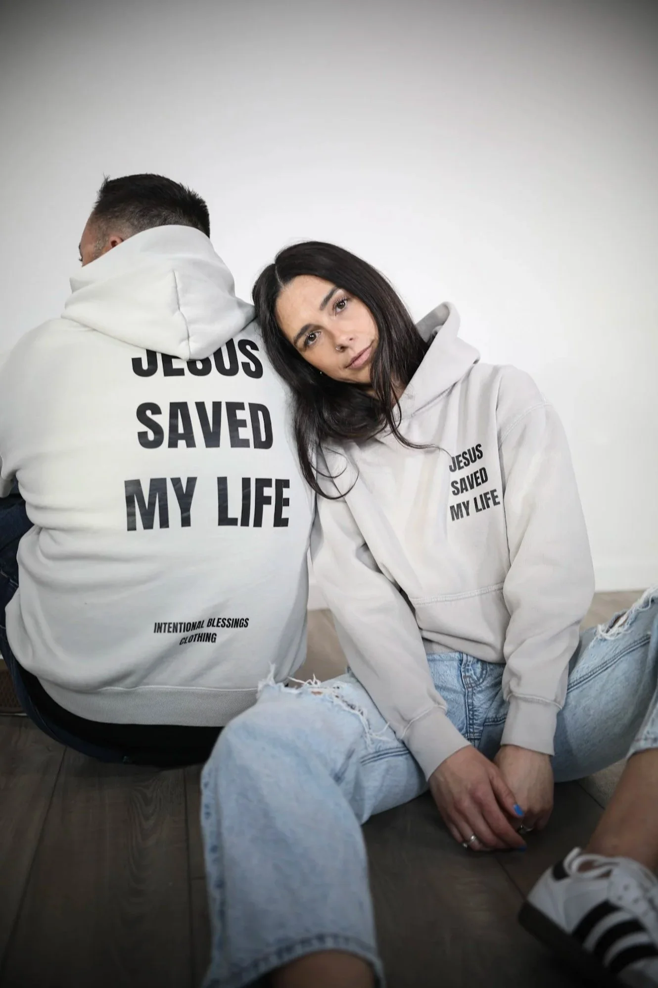 A woman with long dark hair and light skin is sitting on the floor, slightly leaning her head on the shoulder of a man with light skin and short dark hair, who is turned away. Both are wearing white hoodies with black text that reads 'JESUS SAVED MY LIFE'. The woman is looking directly at the camera with a gentle expression, and they are both casually dressed, with the woman in ripped jeans and sneakers. The background is plain and light-colored.