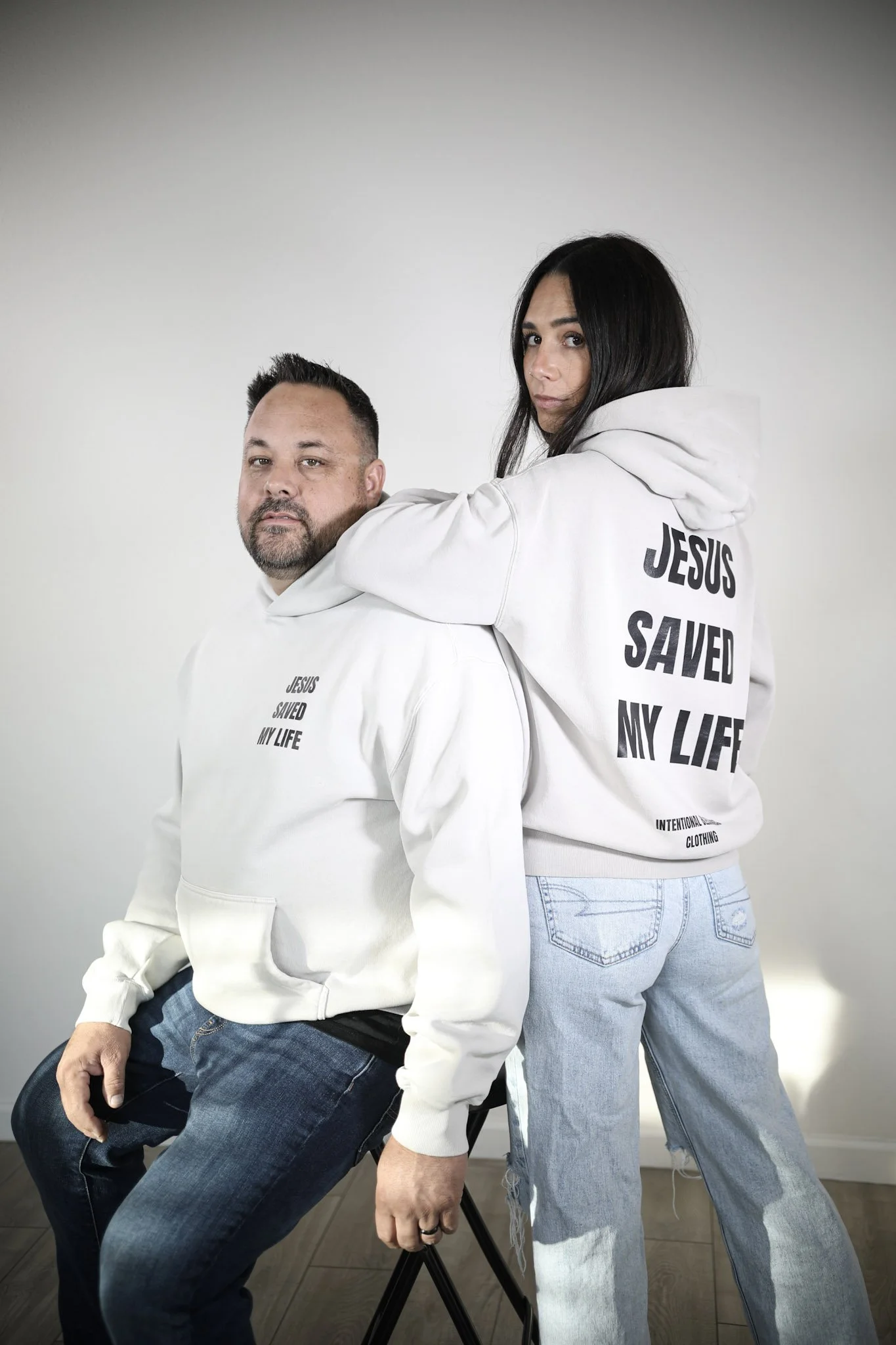 A man sitting on a chair and a woman standing behind him with her arm around his shoulders. Both are wearing white hoodies with the text "JESUS SAVED MY LIFE" printed on the back of the woman's hoodie. The woman is wearing light blue jeans, and the man is wearing darker jeans. They have serious expressions and are in a room with a plain, light-colored wall.