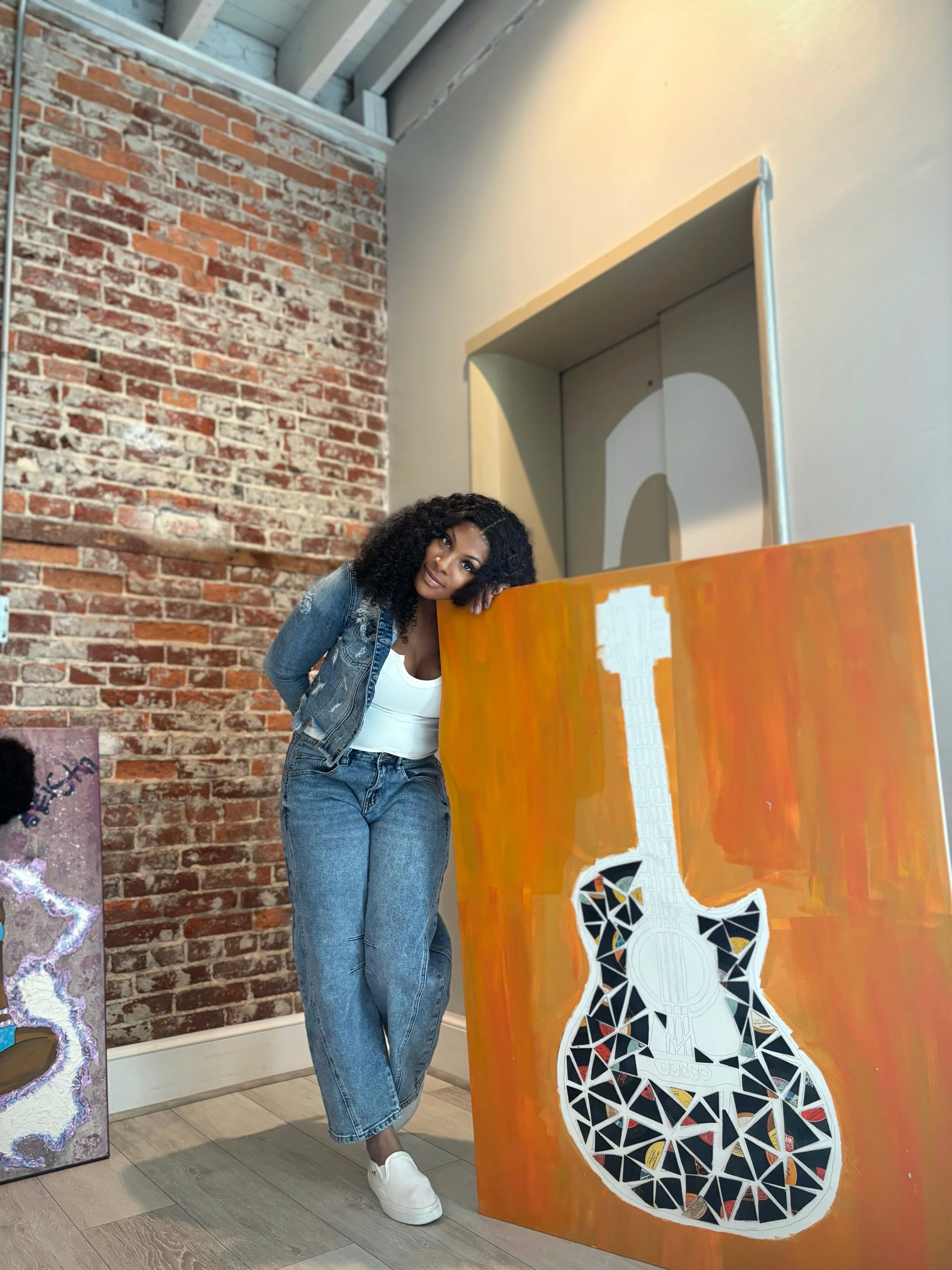 A woman with curly black hair, wearing a denim jacket, white tank top, and jeans, leans her head on a large orange painting of a guitar against a brick wall inside an art gallery.