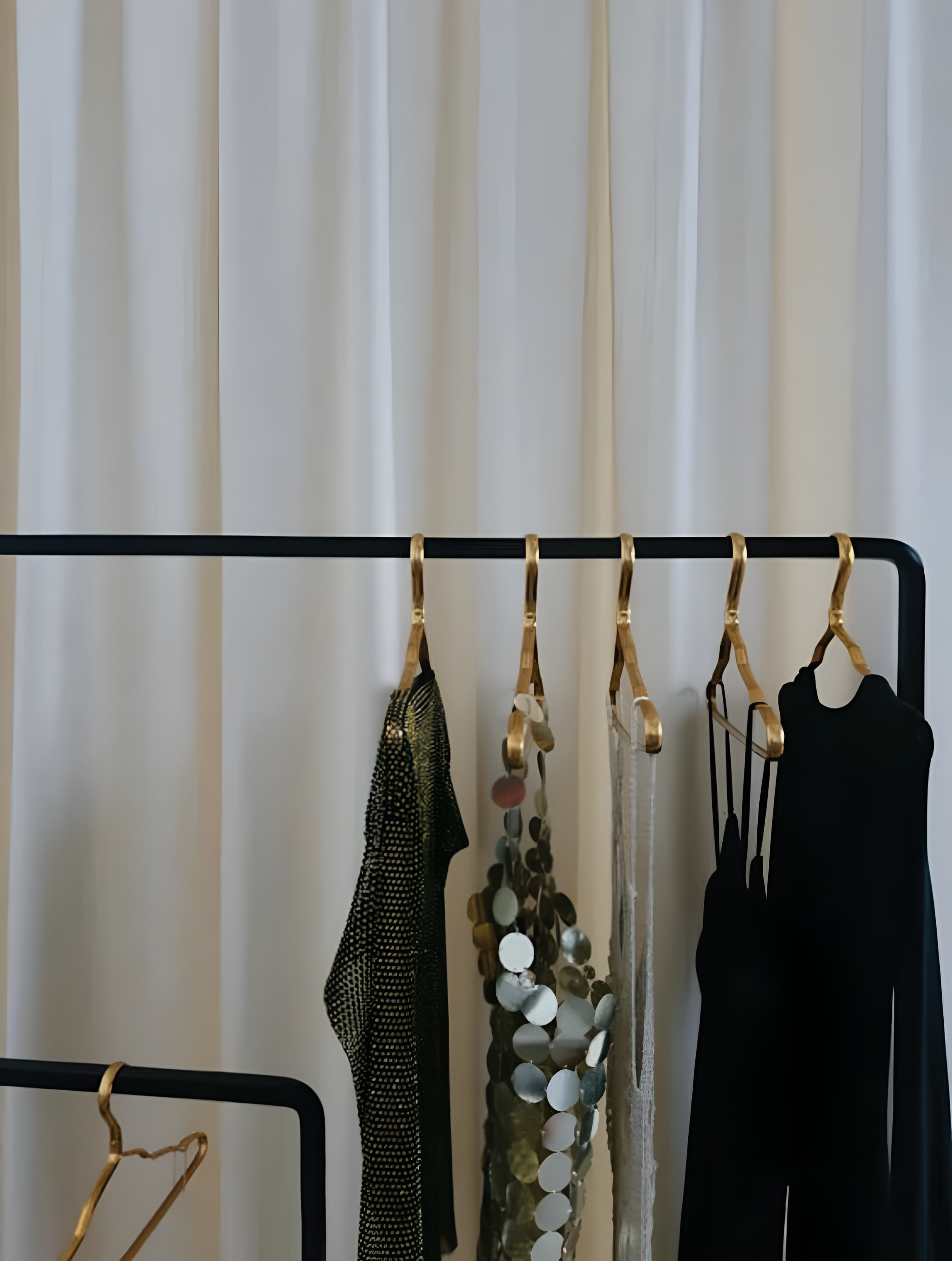Clothing rack with gold hooks holding various black and metallic garments and accessories against a pale curtain background.