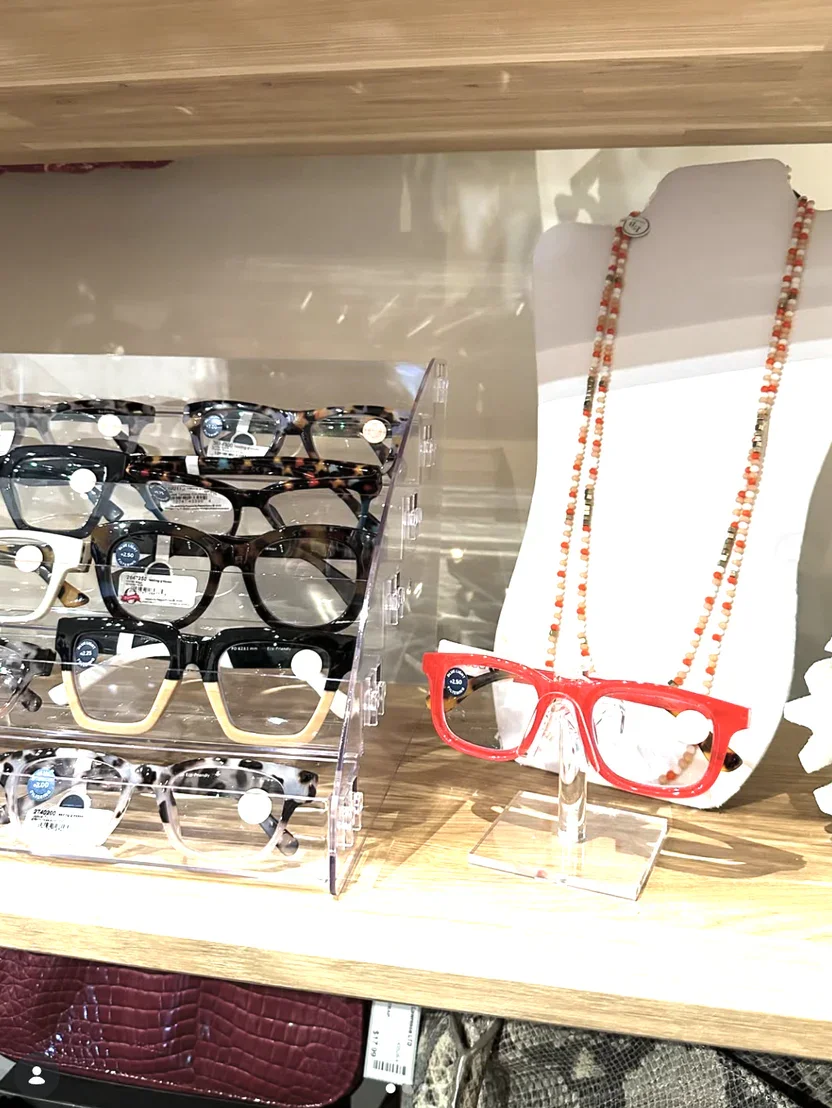 Display of fashionable glasses on a wooden shelf, including a red pair with a plastic frame and a layered store display of various stylish eyewear.
