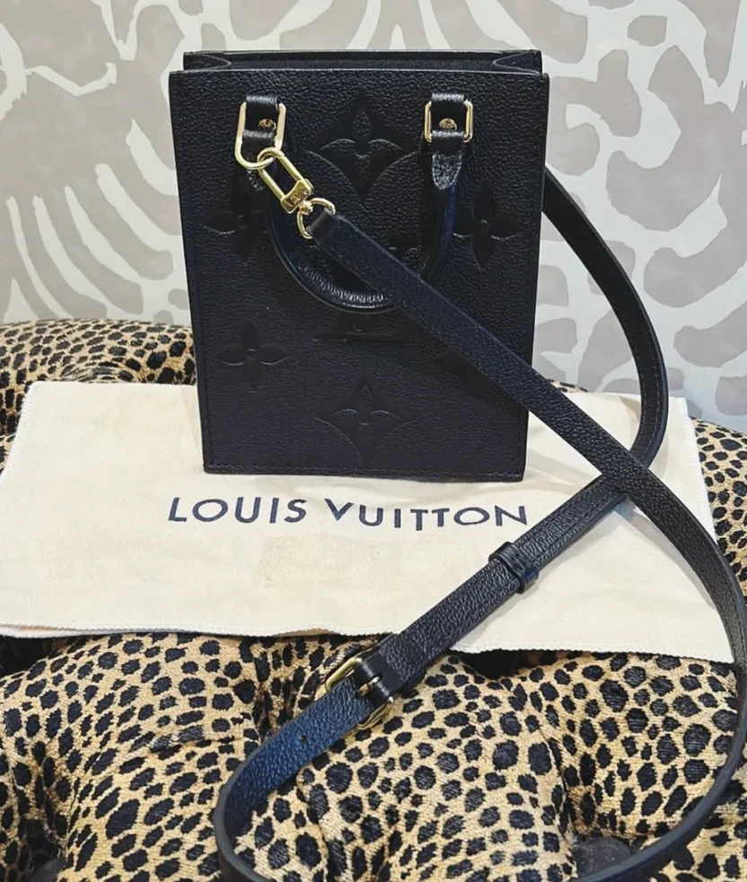 A black Louis Vuitton handbag with embossed monogram pattern, placed on a Louis Vuitton dust bag. The handbag has a shoulder strap and gold hardware, with a leopard print fabric underneath.