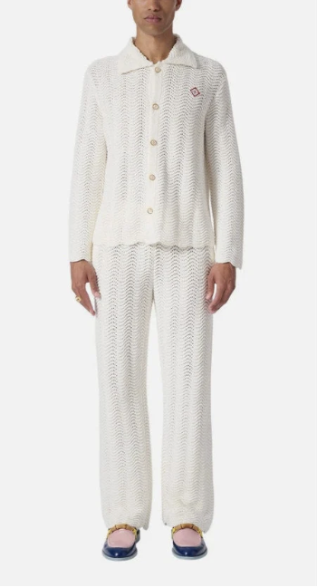 A woman wearing a white knitted cardigan and matching knit pants, standing with her hands by her sides, in blue and yellow slippers.