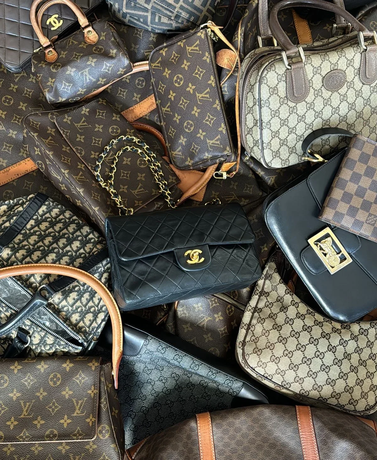 A pile of luxury designer handbags, including Louis Vuitton, Chanel, and Gucci, in various colors and styles.