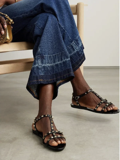 A person sitting on a wooden bench wearing wide-leg denim jeans and black strappy sandals with gold accents, with a clear floor and plain background.