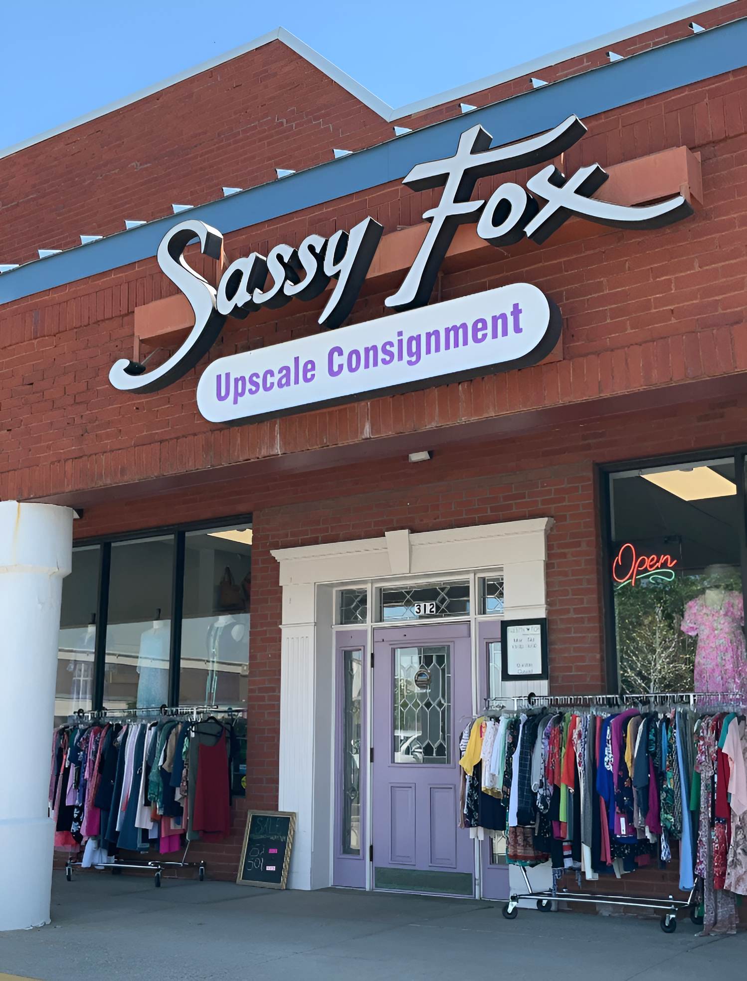 storefront of Sassy Fox Upscale Consignment, with clothing racks outside, a purple door, and an 'Open' sign in the window.
