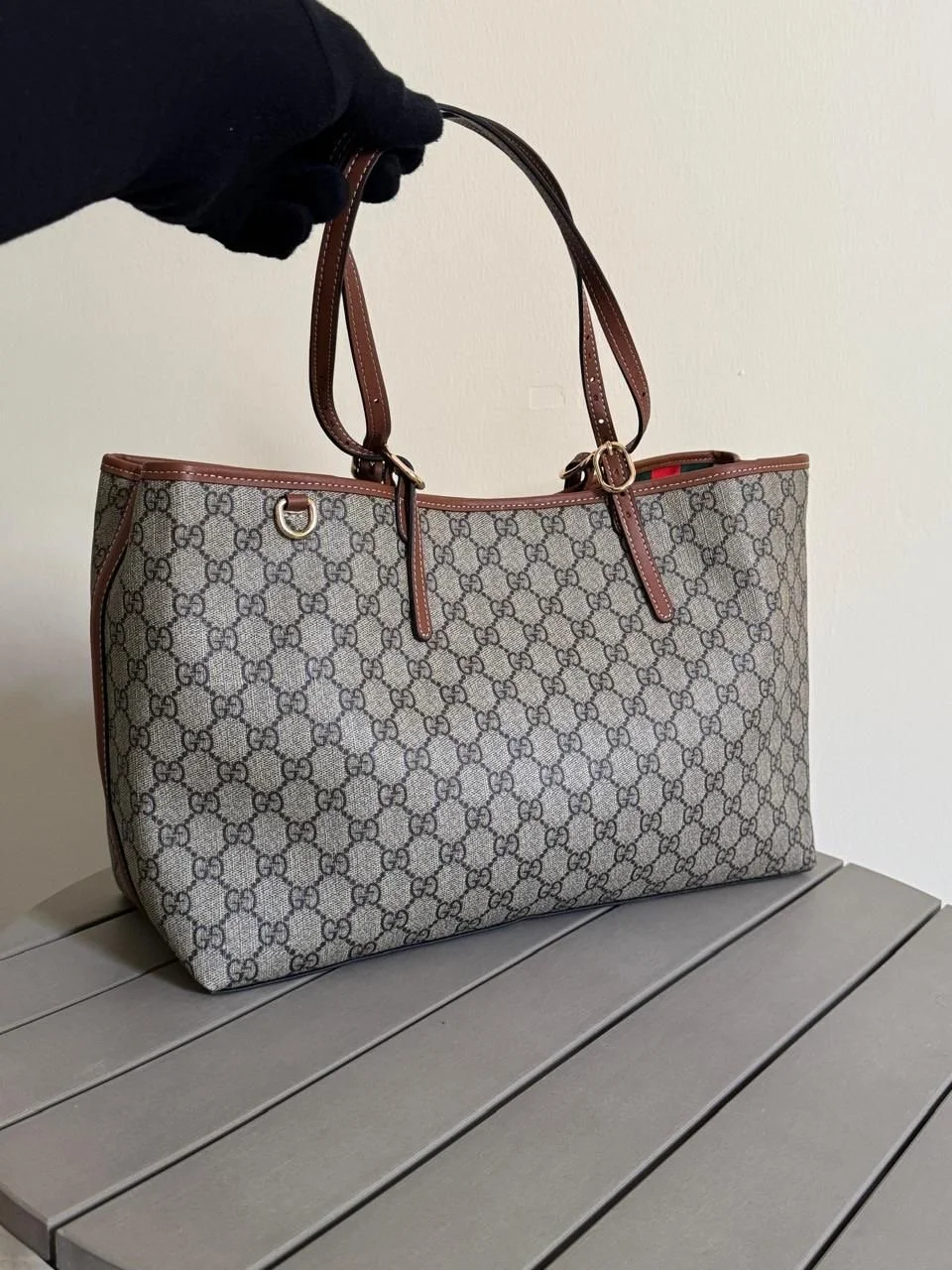 Person in black glove holding a designer tote bag with a beige and brown GG pattern, brown leather handles, and trim, placed on a gray slatted surface against a plain wall.