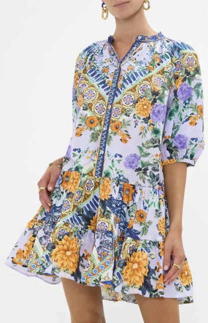 A woman wearing a colorful, floral, bohemian-style dress with yellow, purple, and orange flowers, and intricate pattern details, standing against a plain background.