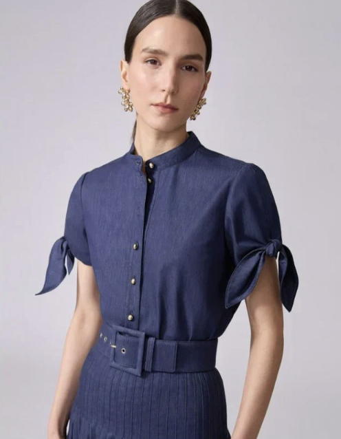 A woman wearing a navy blue shirt with bow-tied sleeves and gold buttons, matching a high-waisted skirt with a belt.