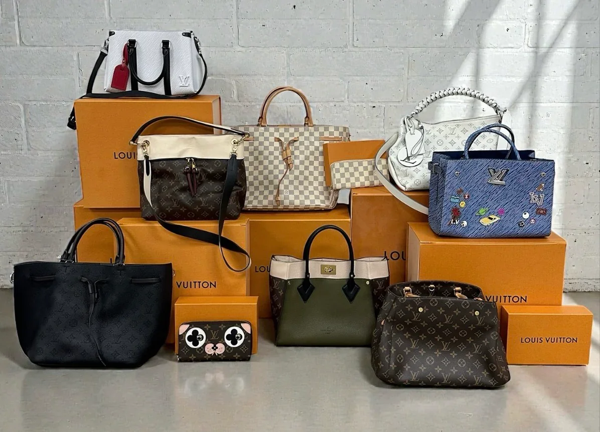 Collection of luxury handbags and wallets displayed on orange Louis Vuitton boxes against a white brick wall.