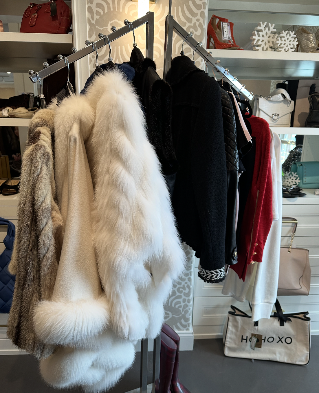 Clothing rack with fur coats, jackets, and sweaters in a boutique store, with handbags and decorative snowflakes on shelves in the background.