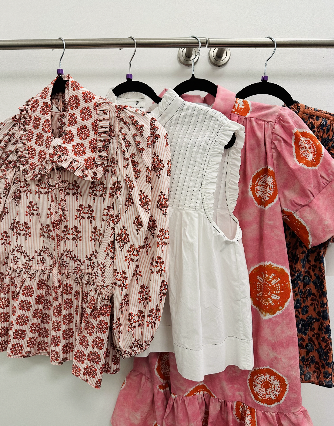 Four colorful dresses hanging on a metal rod. The dresses feature floral and abstract patterns in red, white, pink, and black colors.