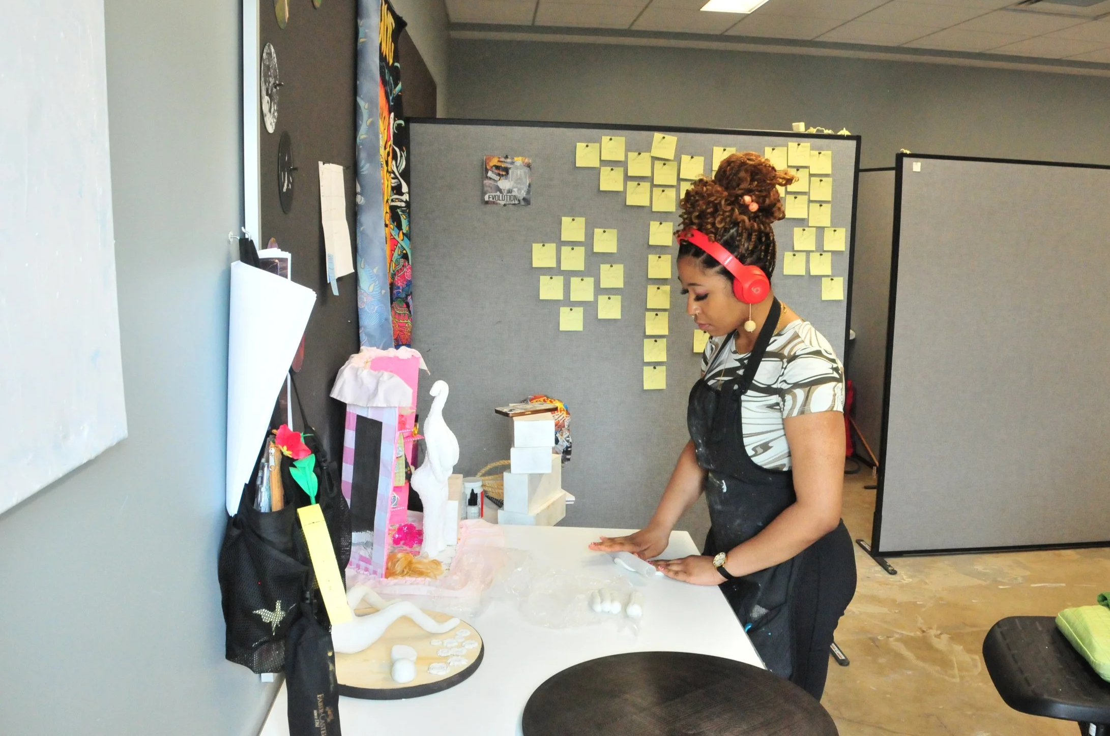 Woman with curly hair wearing red headphones and a black apron working on a craft project at a white table in an office environment.