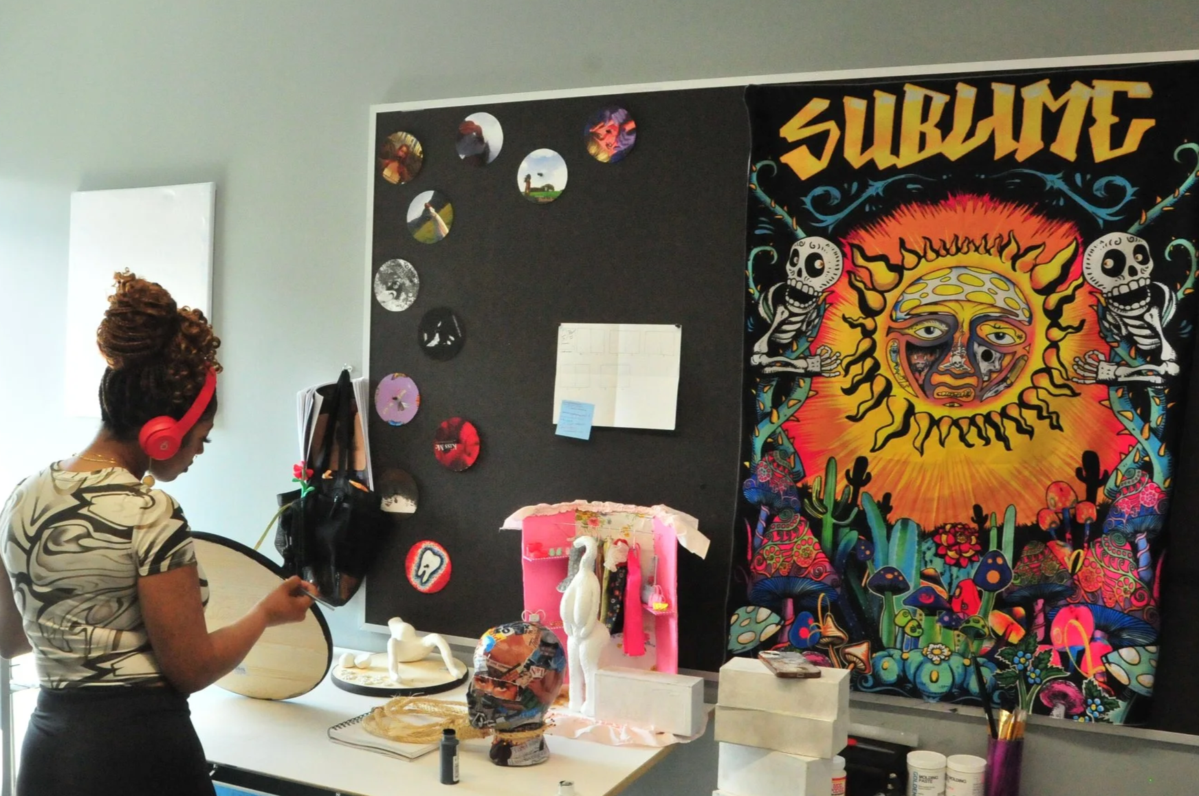 A woman with red headphones standing in front of a black display board with circular images, a large colorful tapestry with the word 'SUBLIME' and a vibrant sun and skeleton design, white sculpture on a table, and art supplies.