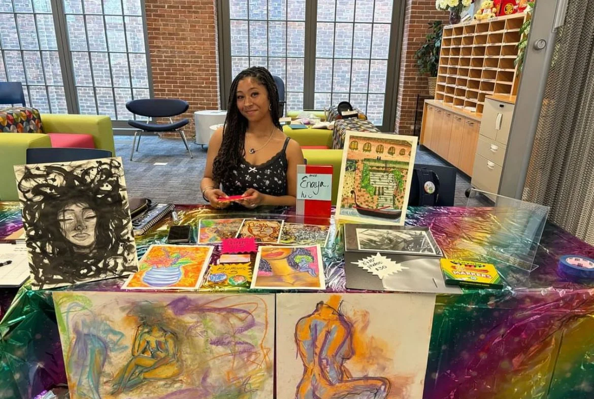 Young woman with long braided hair sitting behind a table at an art fair, displaying various colorful artwork including paintings and drawings, with a sign labeled 'Enaya' on the table, inside a room with large windows and brick walls.