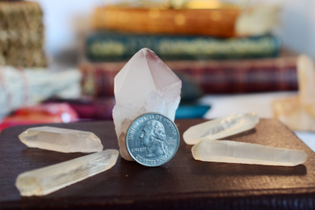 A large clear quartz crystal surrounded by five smaller quartz crystals, with a quarter coin standing upright against the large crystal, on a dark surface with stacked books in the blurred background.