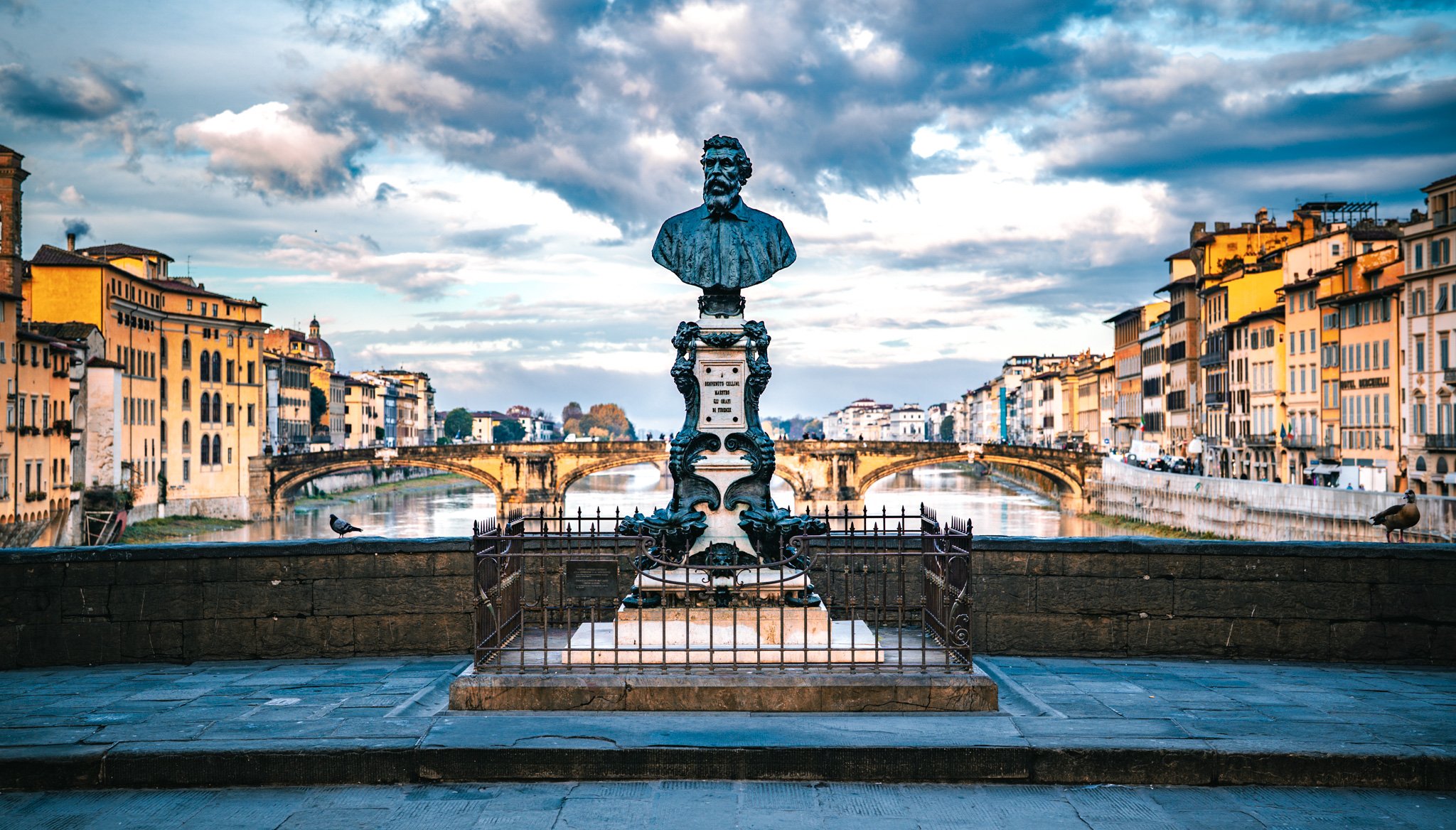 A statue of a man with a beard on a pedestal, overlooking a river with arched bridges and colorful buildings in a European city, under a cloudy sky.