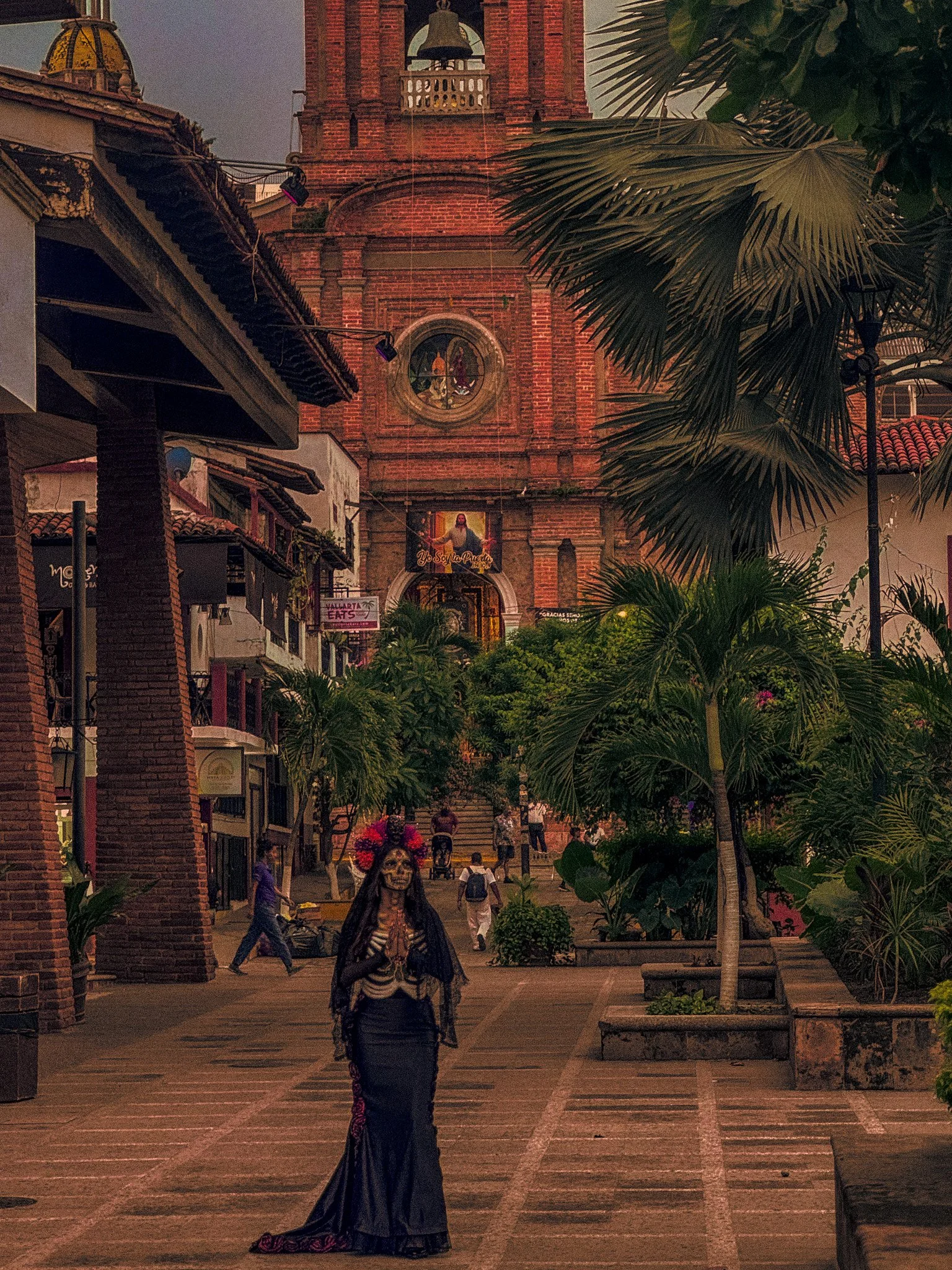 A woman dressed in traditional Mexican clothing, with religious face makeup, walking in a southwestern-style city square with palm trees, brick buildings, and a church in the background.
