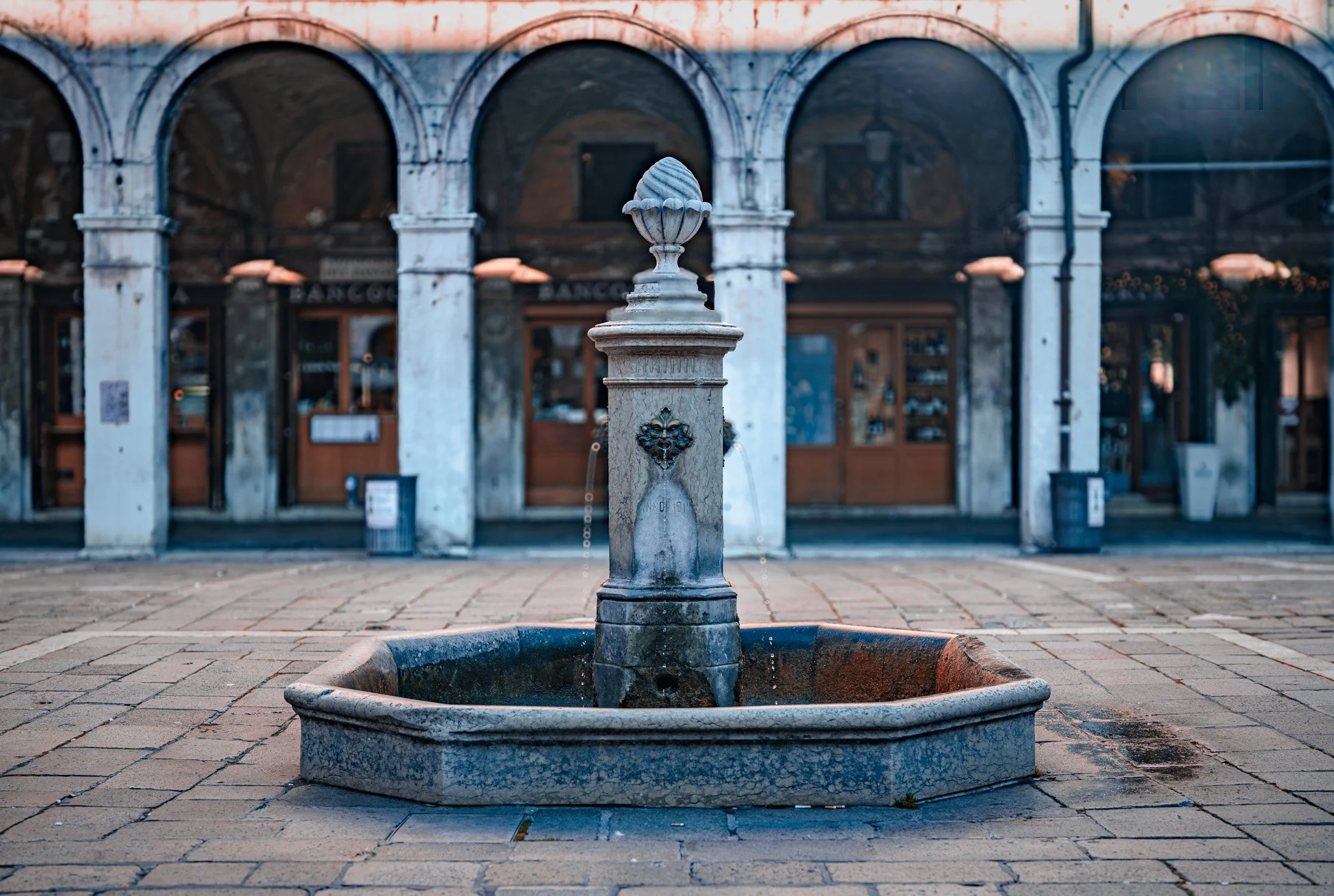 A stone fountain with a classic design in a town square, with an arched building in the background.
