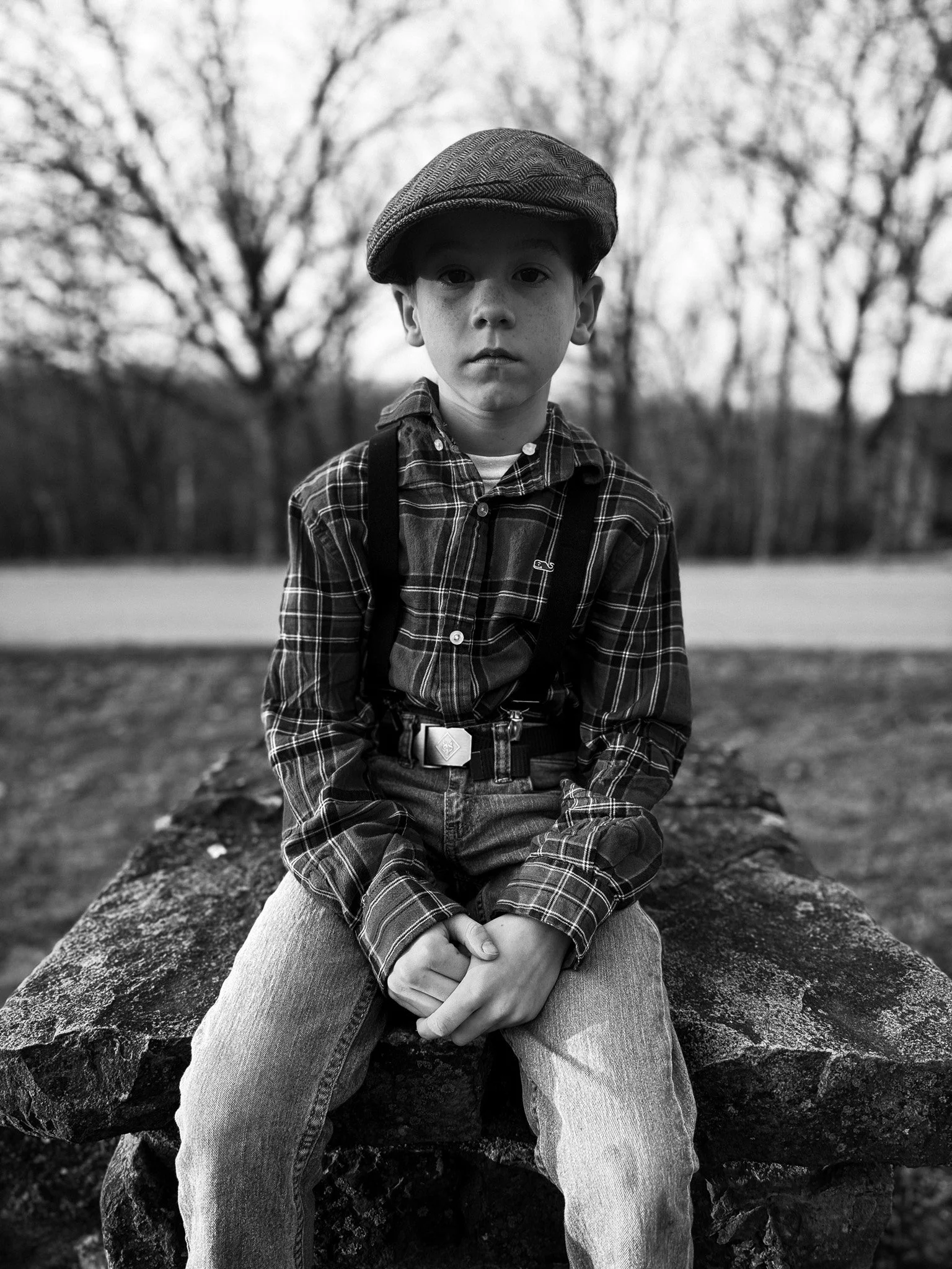 A young boy wearing a flat cap, plaid shirt, suspenders, and jeans, sitting on a large rock outdoors with trees in the background.