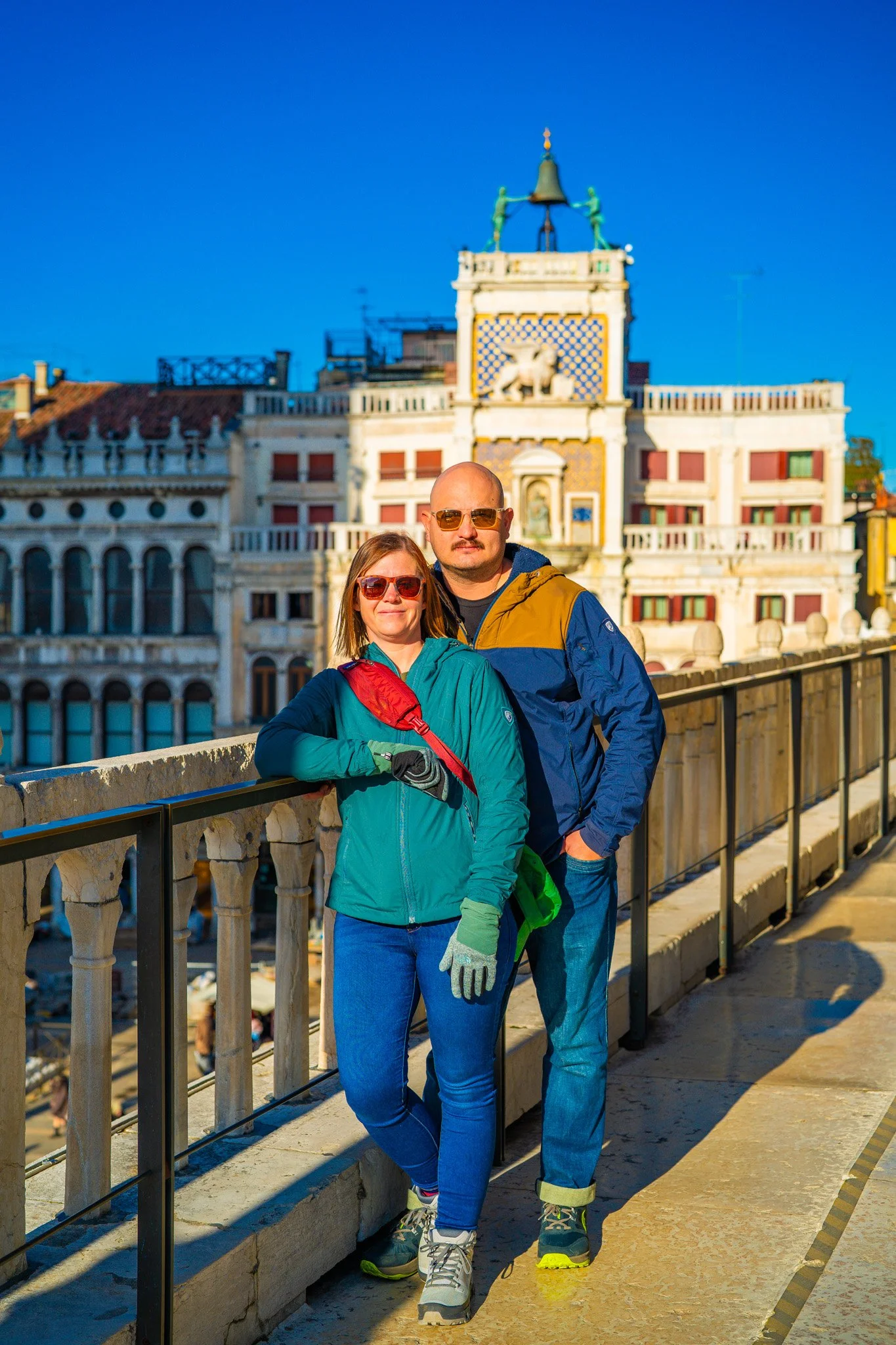 A man and a woman standing on a balcony with historic European buildings behind them, wearing outdoor jackets and sunglasses.