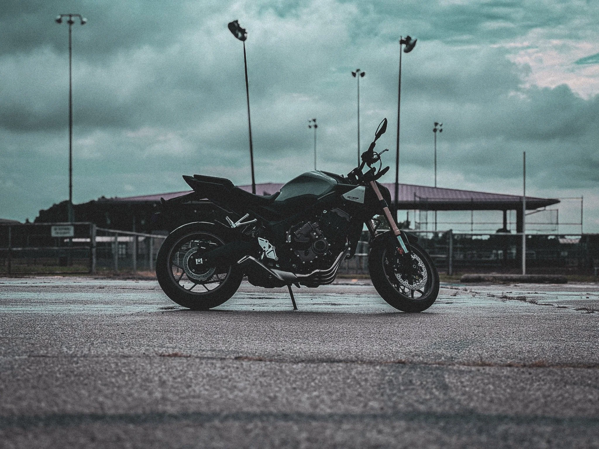 A black motorcycle parked on a concrete surface outdoors with a cloudy sky overhead and a fenced area in the background.