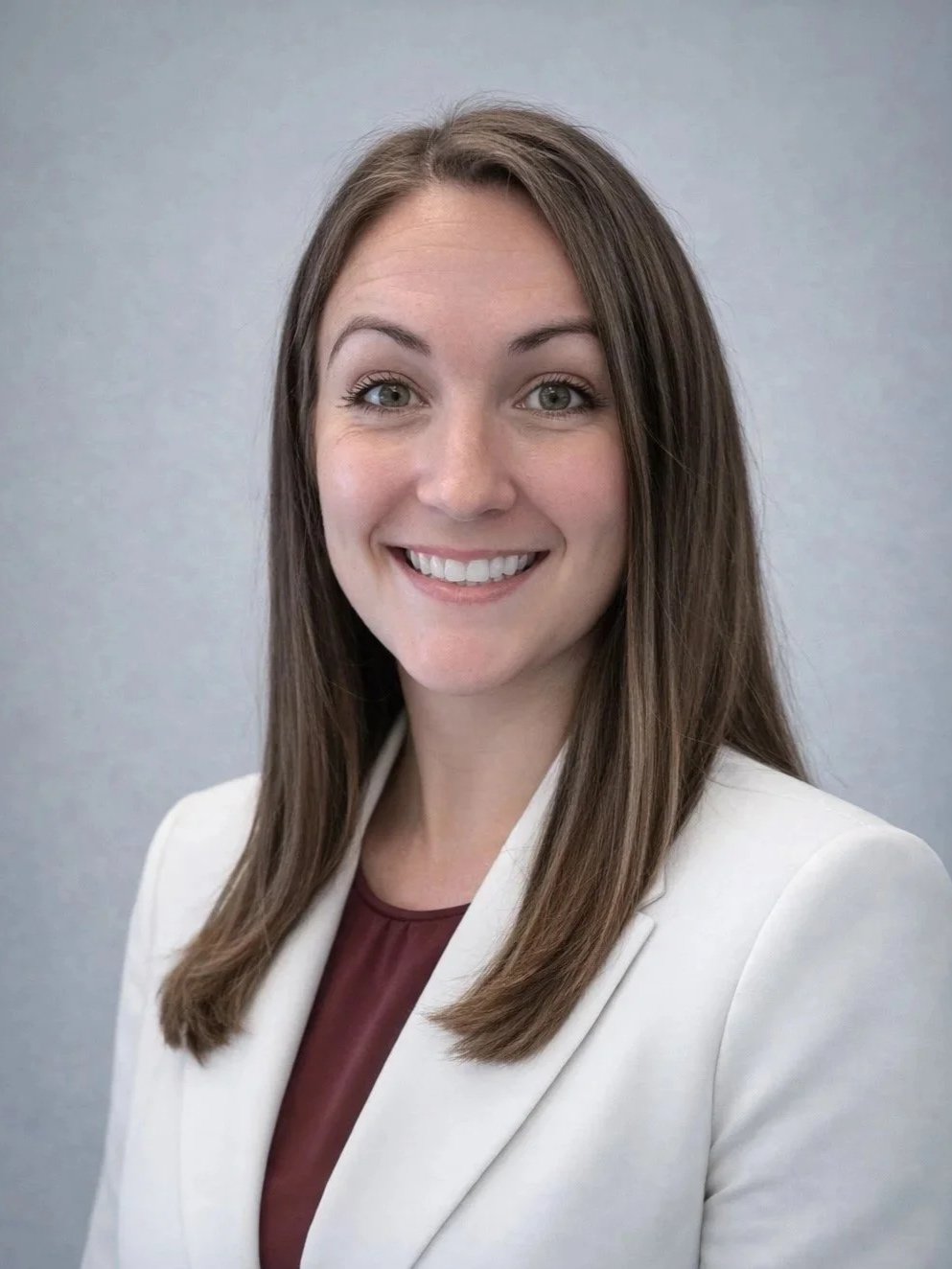 A woman with long brown hair smiling at the camera, wearing a white blazer and maroon top against a plain gray background.