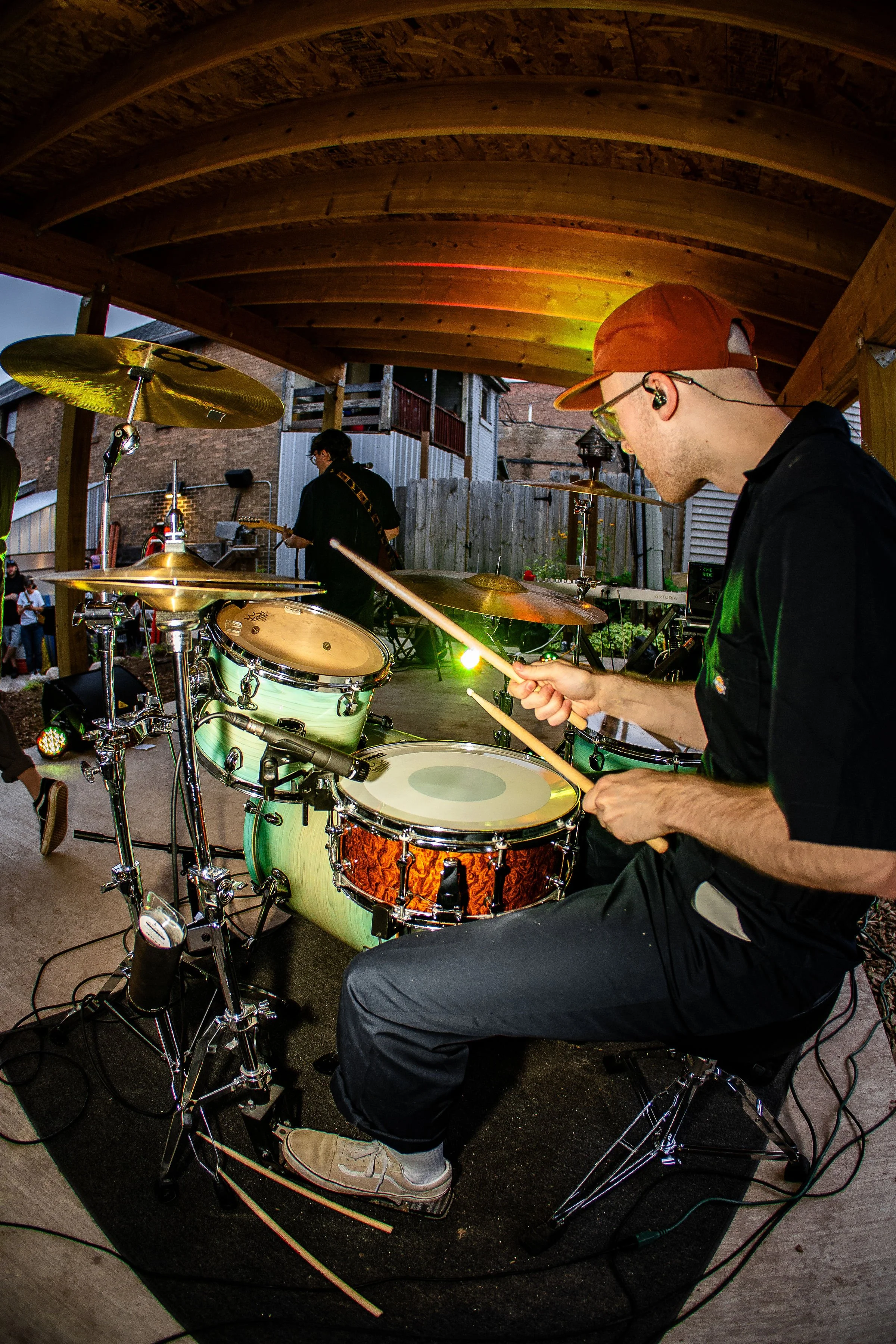 A person playing a drum set at an outdoor concert, with another musician in the background, under a wooden pavilion.