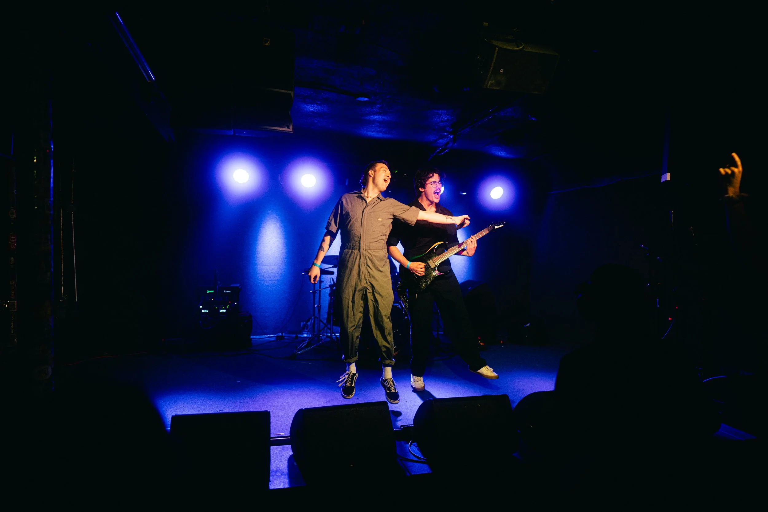 Two performers on stage under blue lights, one singing and the other playing guitar, with dark surroundings and audience silhouettes in front.