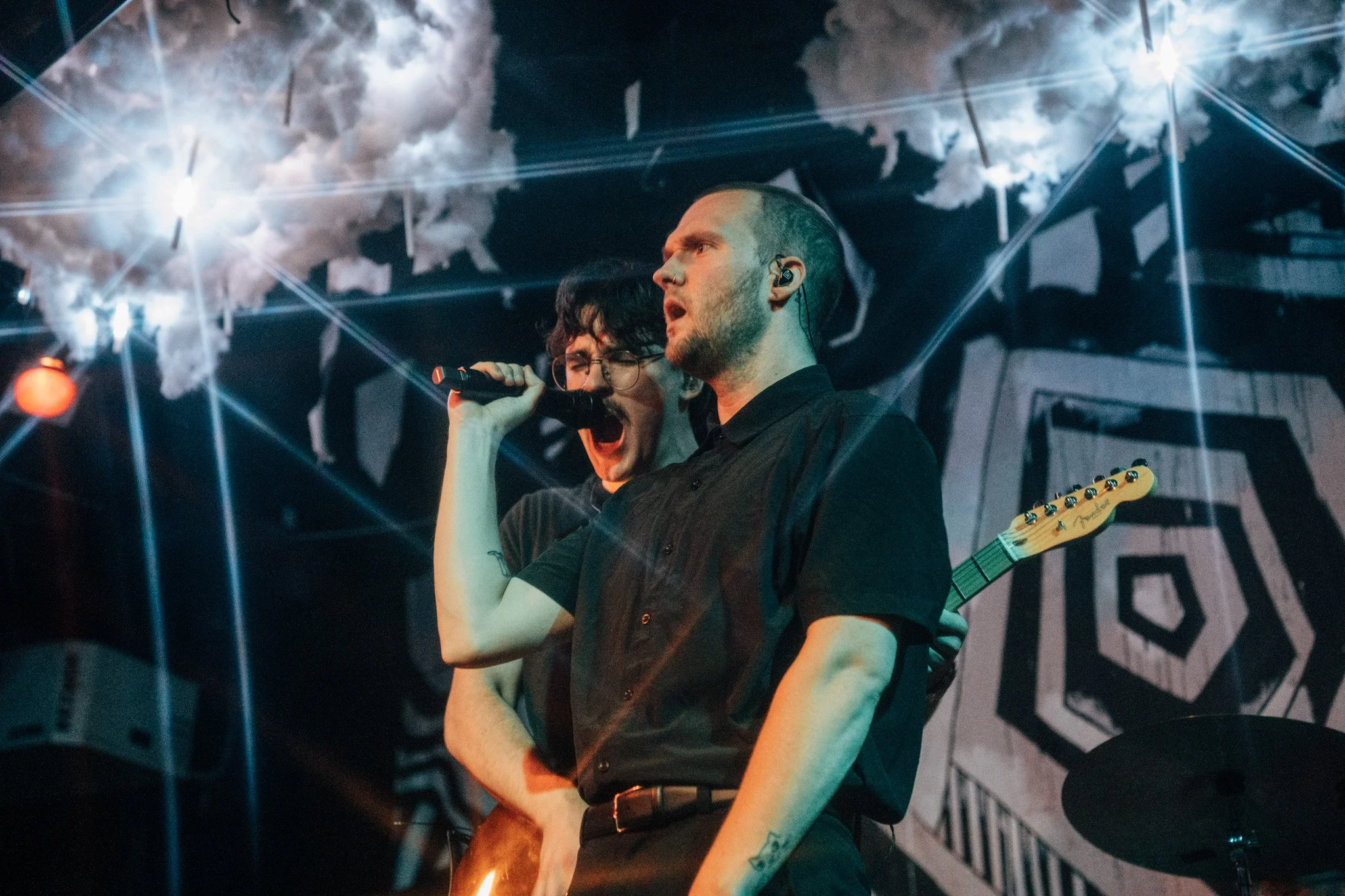Two musicians performing on stage, one with a microphone and the other with a guitar, under theatrical lighting and smoke effects.