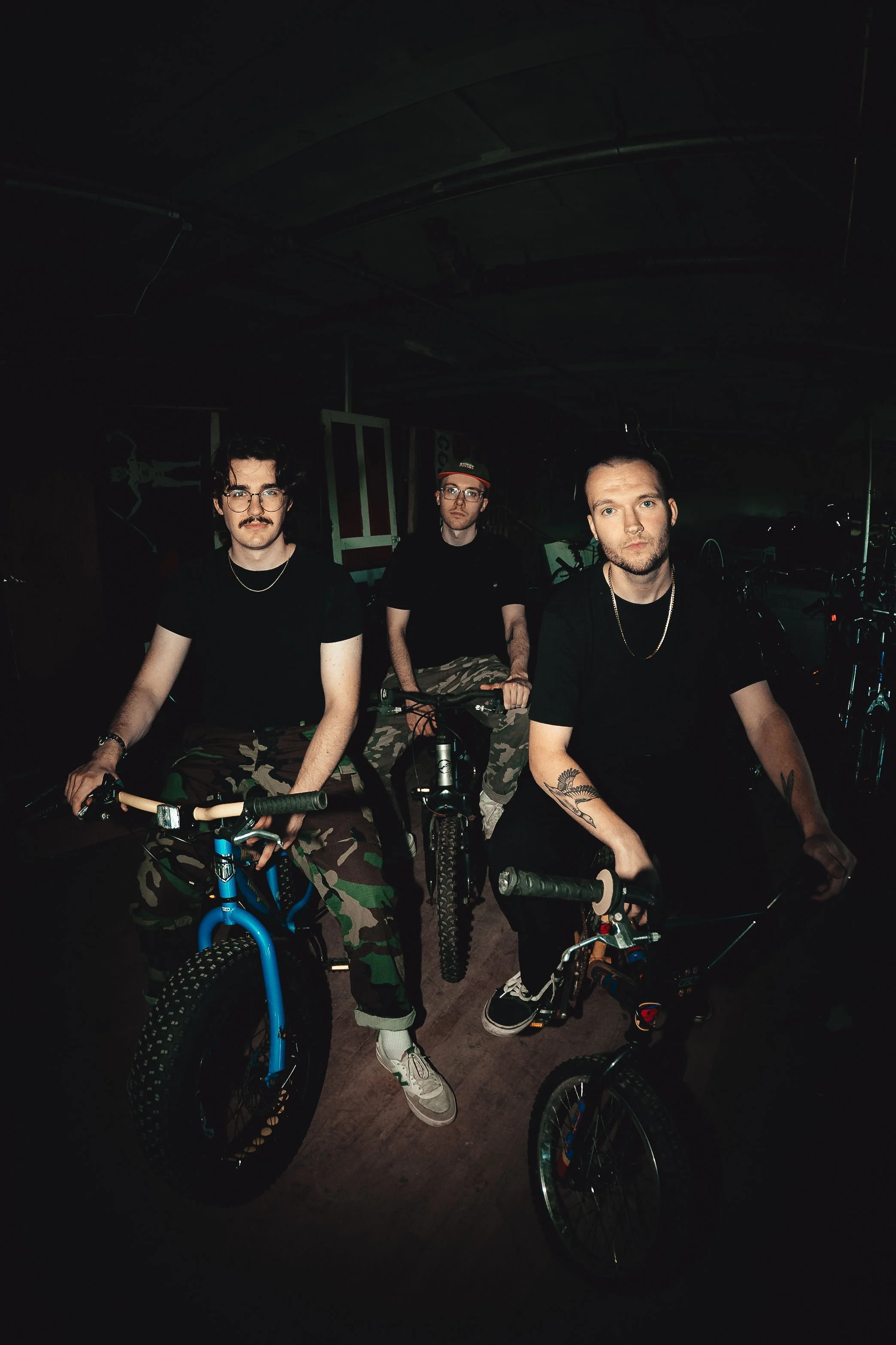 Three young men with bicycles in a dimly lit indoor space, dressed in black t-shirts and camouflage pants, posing for a photo.